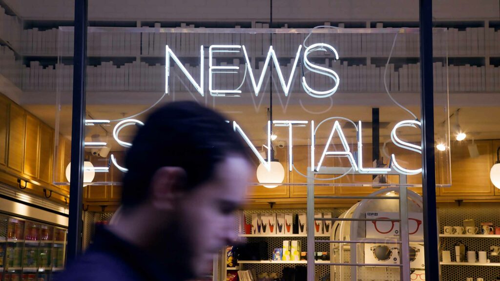 A man walking past a shop window with a neon sign spelling 'News Essentials"